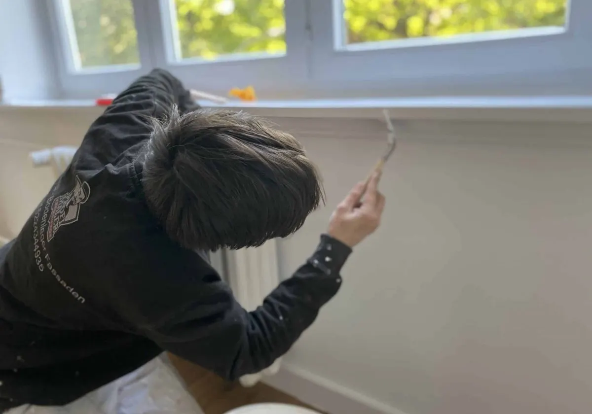 Mitarbeiterin in schwarzer Arbeitsjacke streicht mit dem Pinsel die Wand unter einer Fensterbank