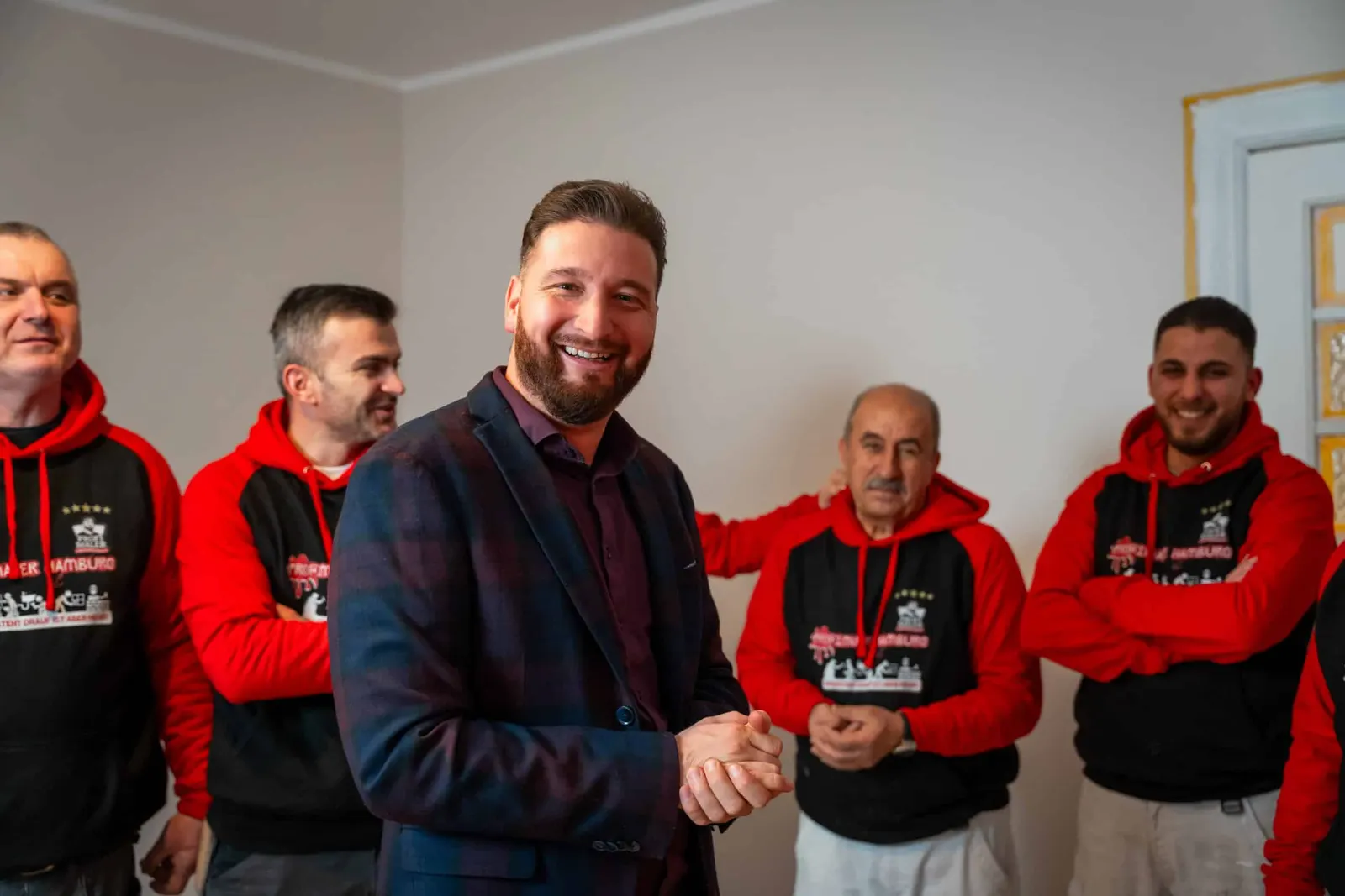 Timo Brandt im karierten Sakko lacht in die Kamera, hinter ihm vier Mitarbeiter in rot-schwarzen Hoodies in einem leeren Zimmer